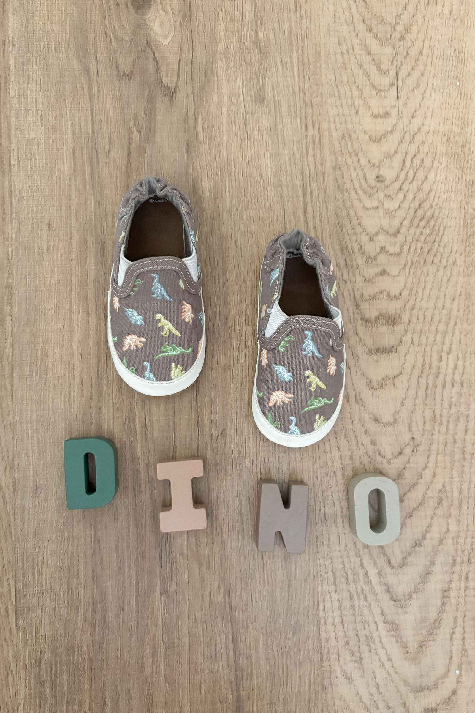 Pair of children's dinosaur shoes on a wooden floor with 'DINO' spelled out below.